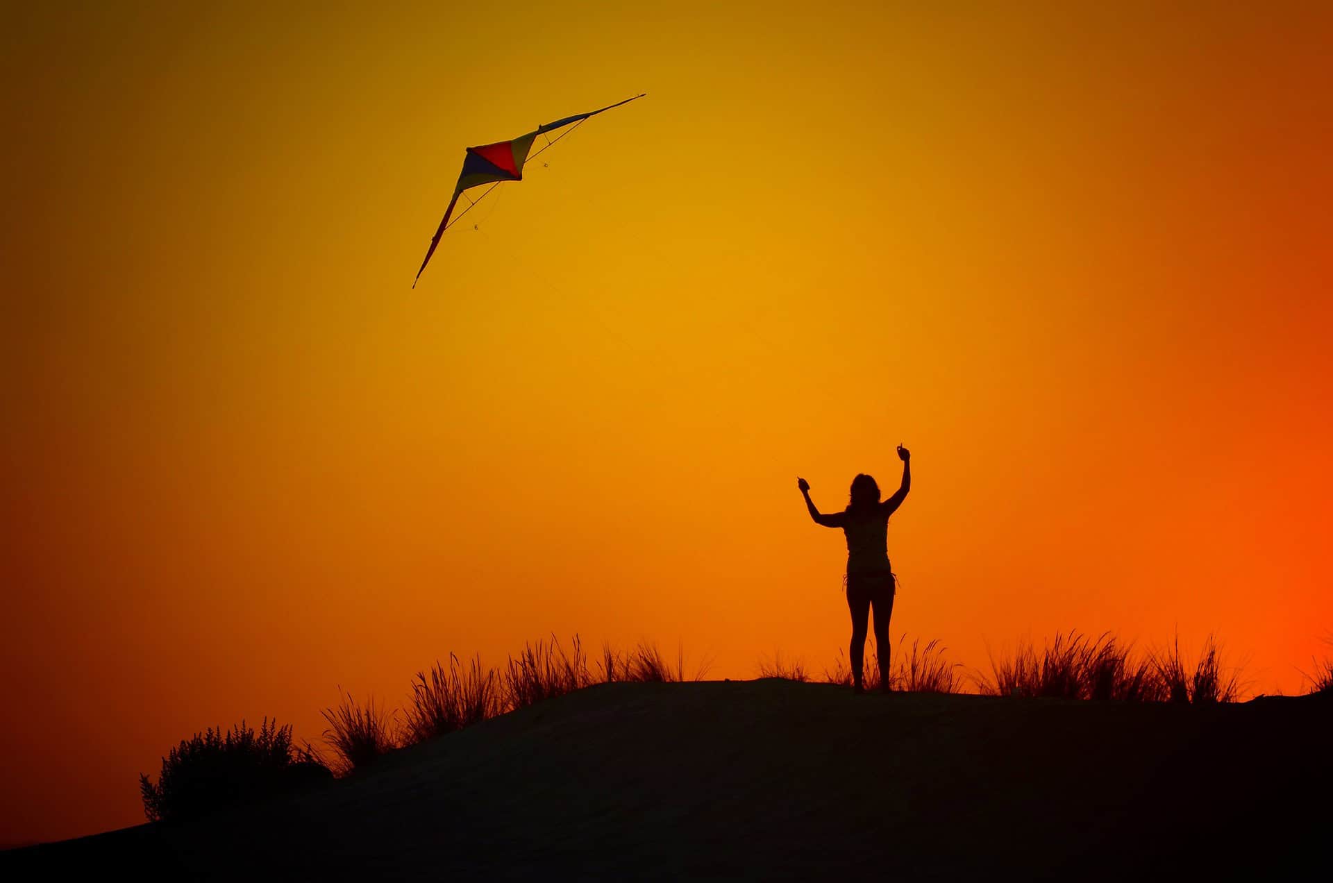 Silhouette of a person with raised arms flying a kite against a vibrant orange sunset.