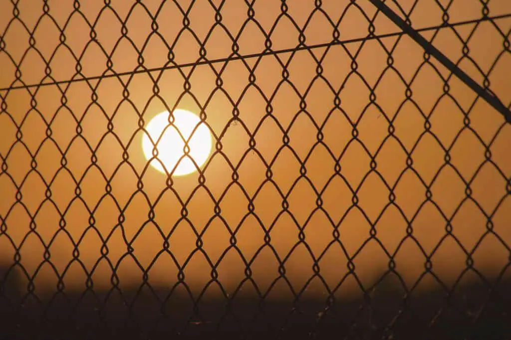 A photograph of the sun setting behind a chain-link fence, casting a warm orange glow across the sky. The intricate diamond-shaped pattern of the fence is silhouetted against the bright, circular sun, creating a striking contrast.